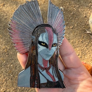 Pode incluir: Um alfinete de esmalte detalhado com uma figura feminina estilizada com pele azul, olhos amarelos e cabelo longo e escuro. O alfinete tem um adorno de cabeça decorativo com detalhes prateados e rosa. A figura usa um top preto e um colar vermelho. O alfinete é segurado em uma mão.