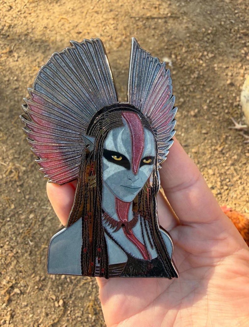 May include: A detailed enamel pin featuring a stylized female figure with blue skin, yellow eyes, and long, dark hair. The pin has a decorative headpiece with silver and pink accents. The figure wears a black top and red necklace. The pin is held in a hand.