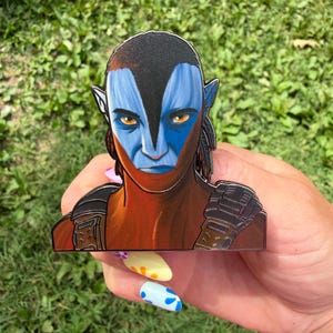 Puede incluir: Un pin de esmalte metálico con un retrato estilizado de un personaje Na'vi de Avatar. La figura tiene piel azul con marcas oscuras, orejas puntiagudas y ojos color ámbar, y lleva piezas de armadura de color marrón oscuro en los hombros.