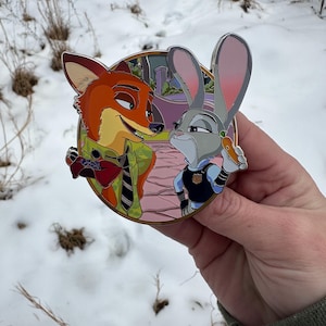 Può includere: Una spilla rotonda e colorata con Nick Wilde e Judy Hopps di Zootropolis. Nick, una volpe, indossa una camicia verde e Judy, un coniglio, indossa un'uniforme della polizia, tenendo in mano una carota. Lo sfondo mostra una scena cittadina.