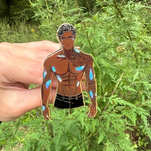 May include: A metal pin depicting a muscular figure with dark skin, black shorts, and a detailed tattoo on the arm. The figure has blue accents on the body and hair. The pin is held against a backdrop of green foliage.