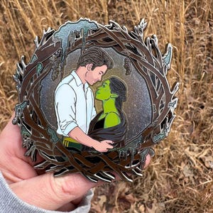 May include: A metal pin featuring a couple embracing, set within a brown, tree-like frame. The woman has green skin and black hair, while the man wears a white shirt. The background is a mix of silver and brown.