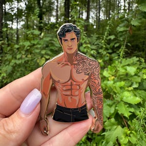 May include: A gold-colored enamel pin featuring a muscular, shirtless figure with a detailed tattoo on his arm. The figure is wearing black pants. The pin is held against a backdrop of green foliage.