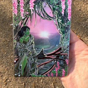 Puede incluir: Un pin rectangular que representa una figura de piel verde con una túnica fluida, sobre un paisaje de puesta de sol. La obra de arte incluye glicinias rosas e intrincado follaje verde, creando una escena de fantasía. El pin se sostiene en una mano.
