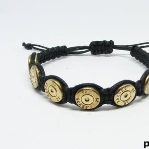 Puede incluir: Una pulsera de cordón negro con una serie de casquillos de bala circulares de latón como elementos decorativos. Los casquillos tienen grabado el texto 7.62x39 21. La pulsera incluye un cierre de nudo deslizante ajustable de cordón negro.