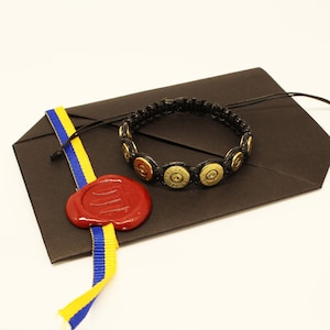 Puede incluir: Un sobre negro con una cinta azul y amarilla y un sello de lacre rojo. Una pulsera de paracord negra con casquillos de bala dorados está encima del sobre.