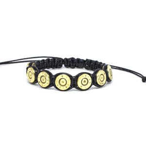 Può includere: Un bracciale in cordino nero con una serie di fondelli di bossoli in ottone disposti in fila. I componenti in ottone sono lucidati e presentano incisioni. Il bracciale è dotato di cordini neri regolabili su entrambe le estremità.