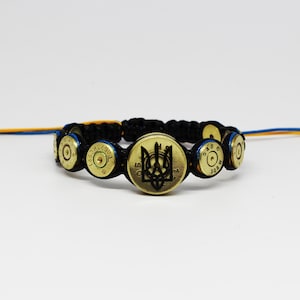 Puede incluir: Una pulsera de cordón negro con una serie de casquillos de bala de latón dispuestos en fila. El casquillo central es más grande y muestra un emblema ucraniano Tryzub en relieve negro. La pulsera termina con cordones ajustables azul y amarillo.