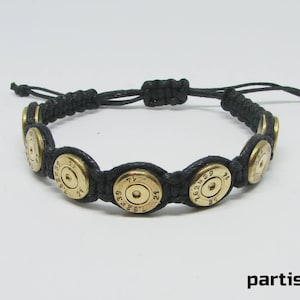 May include: A black cord bracelet featuring a series of circular brass bullet casings as decorative elements. The casings are engraved with the text 7.62x39 and 21. The bracelet has an adjustable sliding knot closure made from the same black cord.