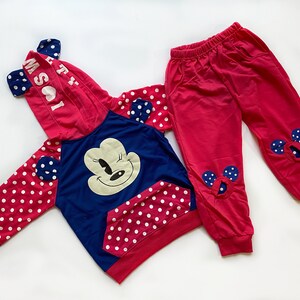 May include: A two-piece children's outfit featuring a red and blue color scheme. The hooded sweatshirt has a large Minnie Mouse graphic on the front, polka dot sleeves, and a hood with attached ears. The matching pants are red with Minnie Mouse ear accents.