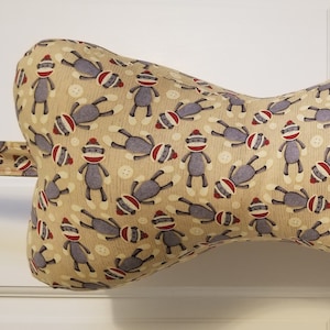 May include: A tan-colored,  bow-shaped neck pillow with a repeating pattern of gray monkeys wearing red hats. The pillow has a tan fabric strap.