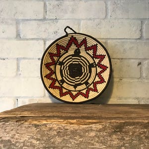 May include: A woven wall hanging with a circular shape and a geometric pattern in shades of brown, white, and red. The hanging has a loop at the top for hanging.