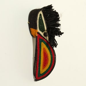 Puede incluir: Una máscara tejida con un diseño negro, verde, amarillo, rojo y blanco. La máscara tiene un pico largo y puntiagudo y una franja de fibras negras en la parte superior.