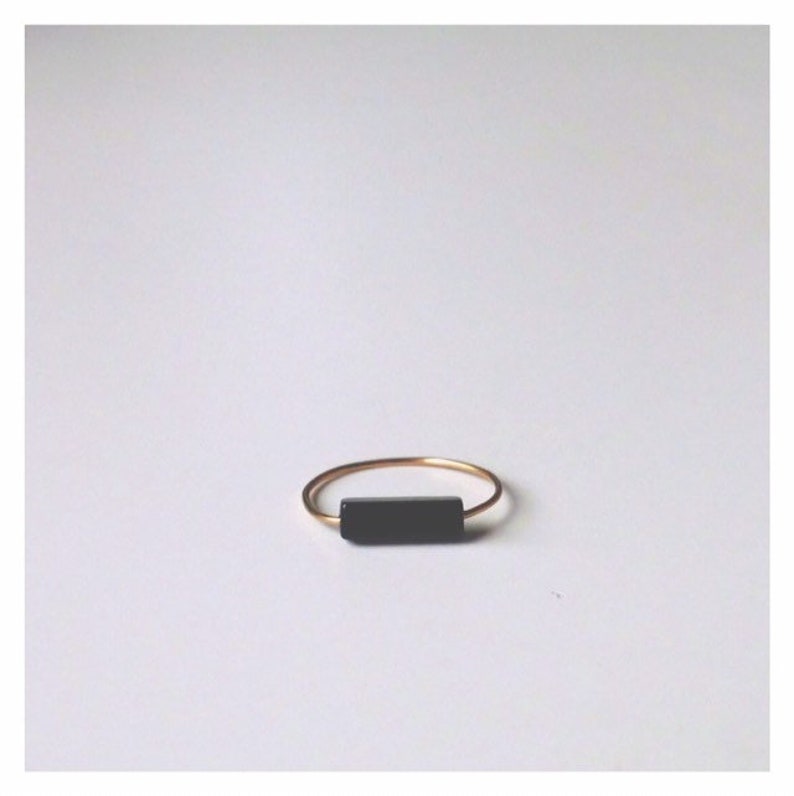 Pu&ograve; includere: Un sottile anello in metallo color oro caratterizzato da un unico inserto a barra rettangolare nera orizzontale centrato sulla fascia. L'anello presenta una finitura liscia e lucida e un design semplice che mette in risalto il contrasto tra oro e nero.