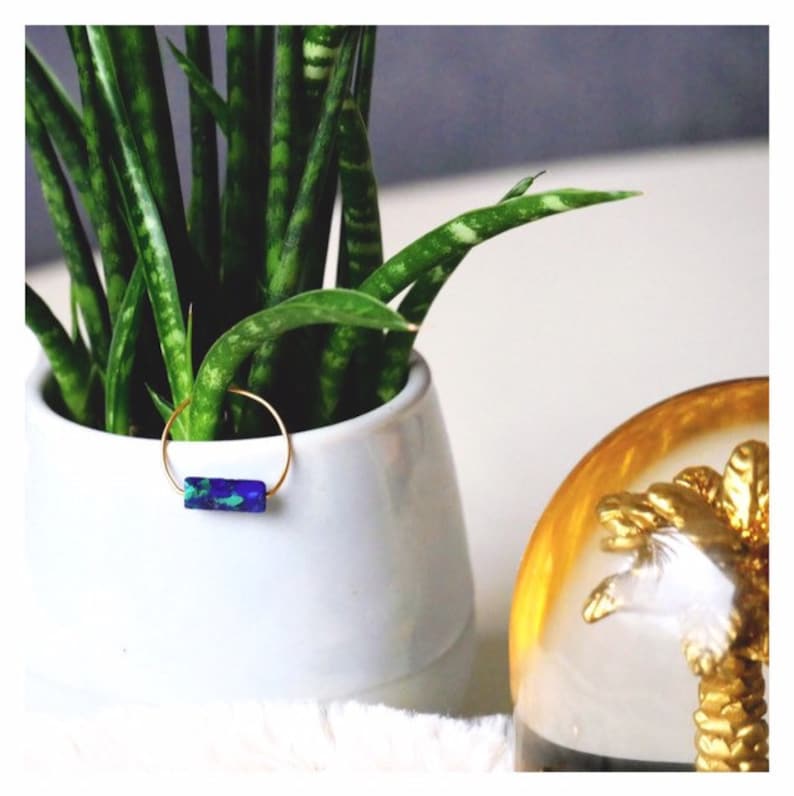 Pu&ograve; includere: Un sottile anello circolare color oro con un inserto rettangolare in pietra iridescente blu e verde. L'anello &egrave; posizionato su una foglia di pianta verde all'interno di un vaso di ceramica bianco, con un oggetto decorativo dorato visibile a destra.