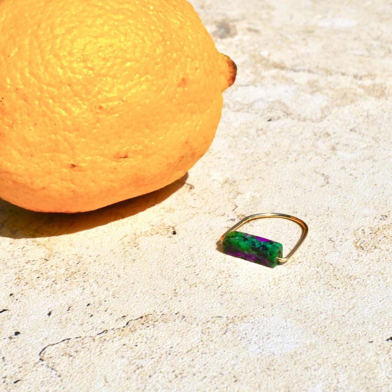 Pu&ograve; includere: Un anello color oro caratterizzato da un inserto a barra rettangolare in pietra screziata verde e viola. L'anello &egrave; posizionato accanto a un limone giallo intero, evidenziando il contrasto tra la fascia metallica e l'inserto in pietra.