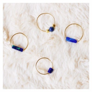 Pu&ograve; includere: Quattro anelli sottili in filo dorato, ognuno caratterizzato da distinti inserti in pietra blu. Un anello presenta una pietra rettangolare blu e verde, un altro una perla blu, uno una barretta blu e il quarto una perla blu con due piccoli accenti dorati.