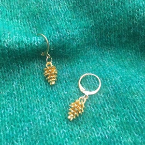 Peut inclure: Une paire de boucles d'oreilles dorées avec un motif de pomme de pin. Les boucles d'oreilles sont suspendues à un anneau.