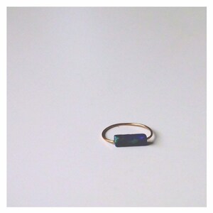 Pu&ograve; includere: Un sottile anello in metallo color oro caratterizzato da una piccola pietra rettangolare blu scuro con macchie iridescenti verdi e blu incastonate orizzontalmente sulla fascia. L'anello ha una struttura semplice in filo circolare sottile.