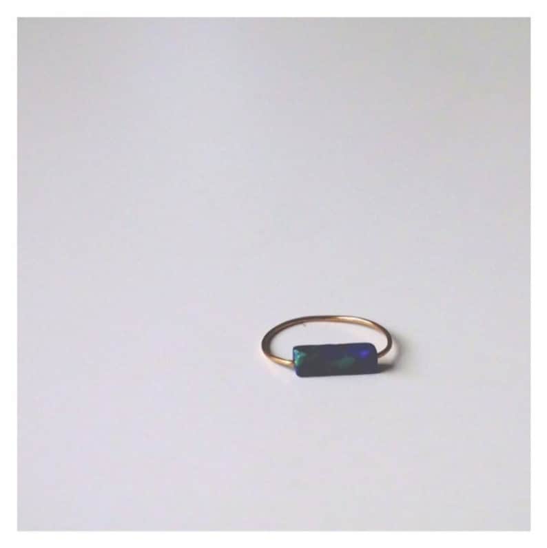 Pu&ograve; includere: Un sottile anello in metallo color oro caratterizzato da una piccola pietra rettangolare blu scuro con macchie iridescenti verdi e blu incastonate orizzontalmente sulla fascia. L'anello ha una struttura semplice in filo circolare sottile.