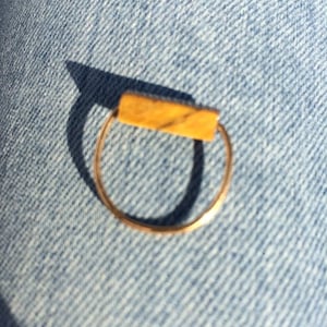 Pu&ograve; includere: Un sottile anello in metallo color oro con un inserto rettangolare orizzontale. La barra presenta una calda tonalit&agrave; giallo-marrone con sottili venature scure sulla superficie, conferendo un aspetto simile alla pietra naturale sulla parte superiore.