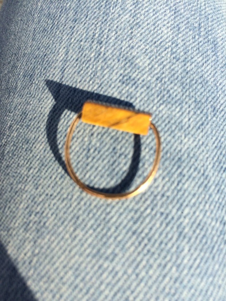 Pu&ograve; includere: Un sottile anello in metallo color oro con un inserto rettangolare orizzontale. La barra presenta una calda tonalit&agrave; giallo-marrone con sottili venature scure sulla superficie, conferendo un aspetto simile alla pietra naturale sulla parte superiore.