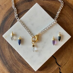 K&ouml;nnte beinhalten: Eine silberne Kettenkette mit einem goldenen Verschluss und f&uuml;nf Kristallspitzenanh&auml;ngern. Die Anh&auml;nger sind klar, blau, lila und schwarz.