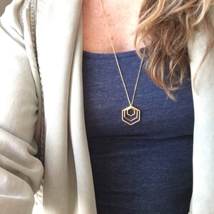 May include: A gold necklace with a geometric pendant featuring three overlapping hexagons. The necklace is worn by a person with long brown hair and a blue tank top.