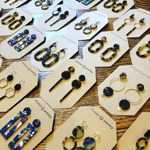 Op de afbeelding: Een assortiment van schildpad- en gepatroneerde hars oorbellen gepresenteerd op bruin karton met de tekst "Inhabit Jewelry" gedrukt op elke kaart. De oorbellen worden getoond in verschillende stijlen, waaronder hoepels, hangers en studs.