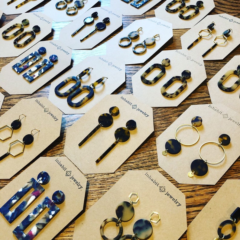 Op de afbeelding: Een assortiment van schildpad- en gepatroneerde hars oorbellen gepresenteerd op bruin karton met de tekst "Inhabit Jewelry" gedrukt op elke kaart. De oorbellen worden getoond in verschillende stijlen, waaronder hoepels, hangers en studs.