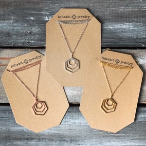 May include: Three gold, silver, and rose gold necklaces with geometric hexagon pendants on brown card displays. Each display has the text "inhabit jewelry" printed on it.