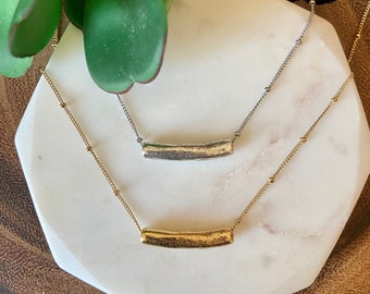Puede incluir: Dos delicados collares con un simple colgante de barra curva. El collar superior tiene un colgante plateado y el collar inferior tiene un colgante dorado. Ambos collares tienen una cadena delicada con pequeñas cuentas.