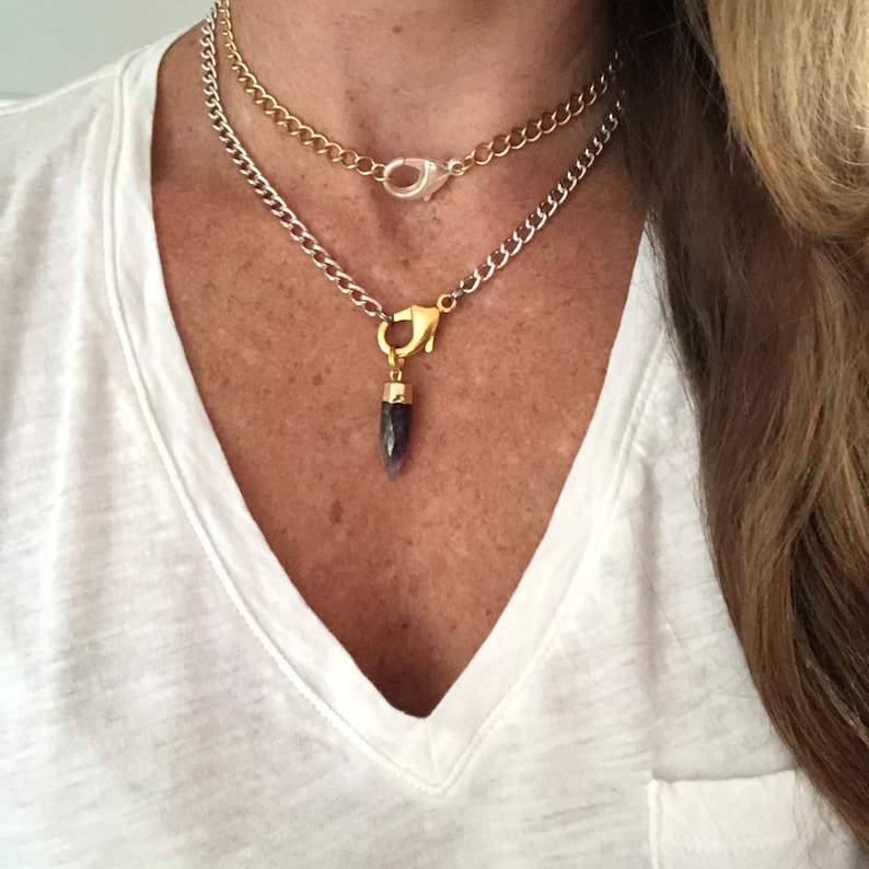 K&ouml;nnte beinhalten: Eine silberne Kettenkette mit einer goldenen Kette dar&uuml;ber gelegt. Die silberne Kette hat einen goldenen Verschluss und einen Anh&auml;nger mit einem dunkelvioletten Edelstein.