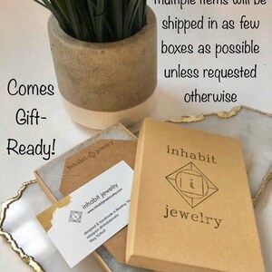 K&ouml;nnte beinhalten: Ein brauner Karton mit dem Aufdruck "inhabit jewelry" auf der Vorderseite. Die Schachtel ist ge&ouml;ffnet und ein kleiner brauner Umschlag mit einer Visitenkarte darin ist sichtbar. Die Visitenkarte tr&auml;gt den Text "inhabit jewelry" und eine Website-Adresse. Der Text "Comes Gift-Ready!" ist auf einer wei&szlig;en Oberfl&auml;che im Hintergrund geschrieben.