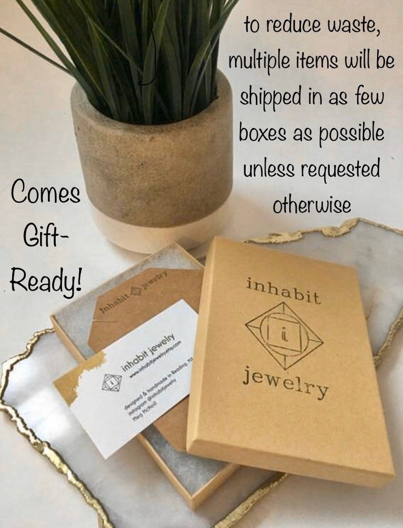 K&ouml;nnte beinhalten: Ein brauner Karton mit dem Aufdruck "inhabit jewelry" auf der Vorderseite. Die Schachtel ist ge&ouml;ffnet und ein kleiner brauner Umschlag mit einer Visitenkarte darin ist sichtbar. Die Visitenkarte tr&auml;gt den Text "inhabit jewelry" und eine Website-Adresse. Der Text "Comes Gift-Ready!" ist auf einer wei&szlig;en Oberfl&auml;che im Hintergrund geschrieben.