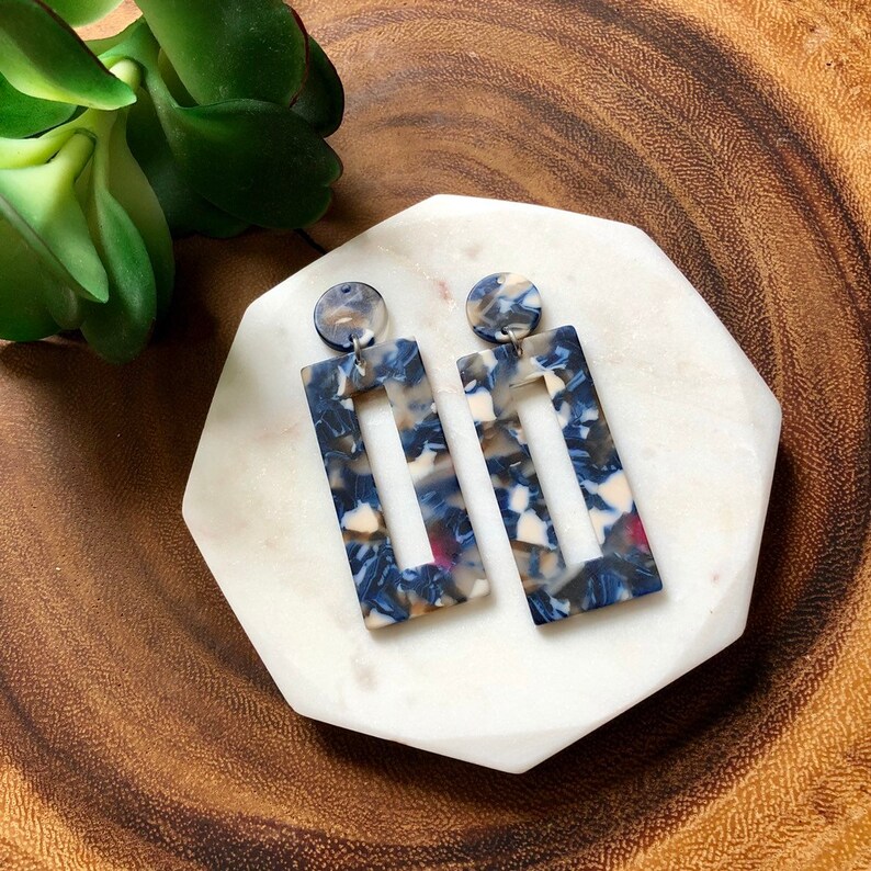 Op de afbeelding: Een paar blauwe, witte en bruine rechthoekige oorbellen met een uitsparing. De oorbellen zijn gemaakt van hars en hebben een marmerlook.