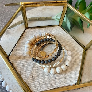 May include: A set of five bracelets featuring a variety of materials and colors. The bracelets include white beads, black lava rock beads, gold-toned metal beads, and a gold-toned metal charm. The bracelets are displayed in a glass hexagonal box.