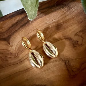 May include: A pair of gold-toned hoop earrings featuring dangling cowrie shell charms. The earrings have a polished metallic finish and a hinged closure mechanism, showcasing the natural ridged texture of the shell design in a bright gold color.