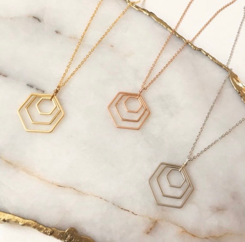 May include: Three gold necklaces with a geometric hexagon pendant. The necklaces are layered on a white marble surface. The hexagon pendant is made of three concentric circles.
