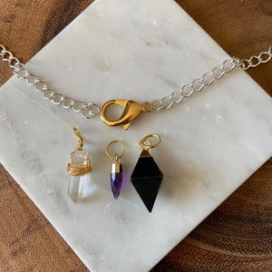 K&ouml;nnte beinhalten: Eine silberne Kettenkette mit einem goldenen Verschluss und vier Kristallanh&auml;ngern. Die Anh&auml;nger sind eine klare Quarzspitze, eine violette Amethystspitze und eine schwarze Obsidianpyramide.