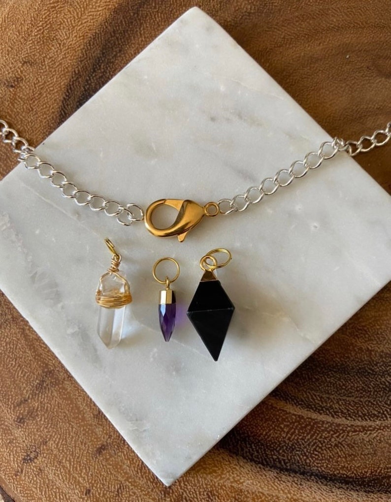 K&ouml;nnte beinhalten: Eine silberne Kettenkette mit einem goldenen Verschluss und vier Kristallanh&auml;ngern. Die Anh&auml;nger sind eine klare Quarzspitze, eine violette Amethystspitze und eine schwarze Obsidianpyramide.