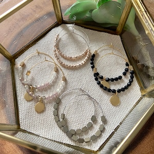 May include: Four pairs of gold hoop earrings with different colored beads. The earrings are displayed in a gold hexagonal box. The beads are pink, black, and gray.