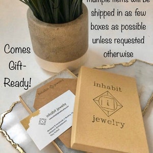 May include: A brown cardboard box with the text "inhabit jewelry" printed on the front. The box is open and a small brown envelope with a business card inside is visible. The business card has the text "inhabit jewelry" and a website address. The text "Comes Gift-Ready!" is written on a white surface in the background.