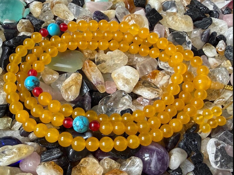 May include: A strand of yellow beads arranged in a circular pattern, featuring interspersed bright turquoise stone beads and small red accent beads. The yellow beads have a smooth, polished finish, while the turquoise beads display dark veining.