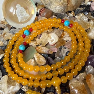 May include: A long strand of yellow beads featuring interspersed bright turquoise and small red accent beads. The necklace includes a larger yellow focal bead and a dangling tassel end, resting on a collection of various polished stones.