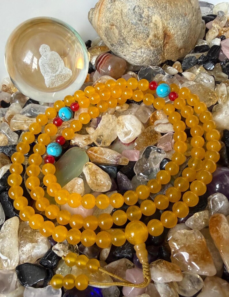 May include: A long strand of yellow beads featuring interspersed bright turquoise and small red accent beads. The necklace includes a larger yellow focal bead and a dangling tassel end, resting on a collection of various polished stones.