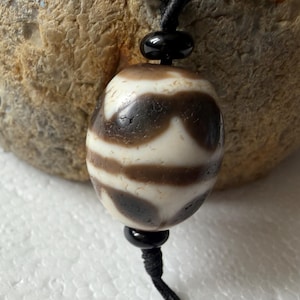 May include: An oval-shaped bead with a white and dark brown patterned surface, featuring a weathered texture. The bead is strung on a black cord and is accented by two small, smooth, glossy black spherical beads positioned above and below it.