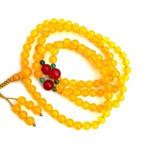 May include: A yellow beaded necklace with a tassel and decorative beads. The necklace features a series of round, yellow beads. Accented with red and green beads, the necklace is displayed against a white background.