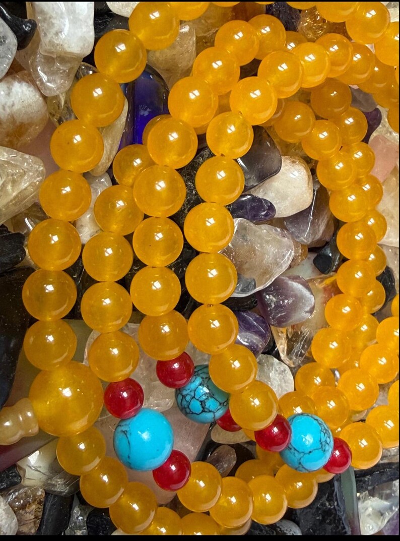 May include: A beaded necklace or bracelet featuring numerous smooth, round, translucent yellow beads. Interspersed among the yellow beads are smaller, vibrant red spherical beads and larger, round, bright turquoise beads with black veining.