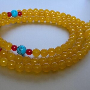 May include: A long strand of prayer beads featuring smooth, translucent yellow beads. The strand is accented with three larger, round, bright turquoise beads with dark veining, each flanked by two smaller, vibrant red spherical beads.